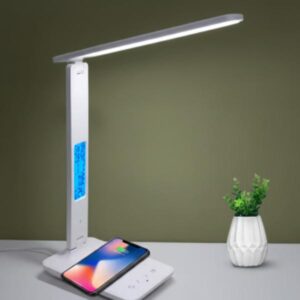 A white LED desk lamp is shown with its light bar illuminated. A smartphone is placed on the base for wireless charging, and a small potted plant in a white vase is positioned next to it. The lamp features a blue digital display on its vertical arm, set against an olive green background