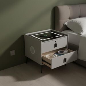 A grey smart bedside table with its bottom drawer partially open, revealing a book and headphones inside, is placed next to a bed with a fabric headboard. The room has neutral-colored walls, showcasing the table's use as functional bedroom furniture.