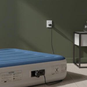 A close-up view of the side of the blue and white inflatable mattress, showing the built-in pump unit plugged into a white wall socket. The wall is painted dark olive green, and a side table with a lamp is partially visible next to the mattress. This highlights the ease of inflation/deflation using the integrated pump.