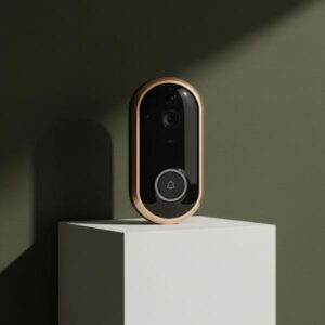 A close-up hero shot of the golden-framed smart video doorbell standing on a clean white cubic block. The background is a solid dark olive green wall with soft shadows, highlighting the product's sleek, rounded design and metallic frame.