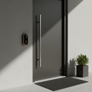 The golden-framed smart video doorbell is installed on a light grey wall next to a modern light grey door with a long stainless steel handle. Sunlight creates a dramatic shadow extending from the corner. A small potted plant adds a touch of greenery to the contemporary entrance.