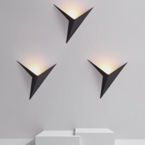 hree black geometric, triangular wall sconces mounted on a light gray or white wall, casting a warm light upwards. The lights are displayed above a minimalist tiered white pedestal, highlighting a clean, modern, and high-contrast design.