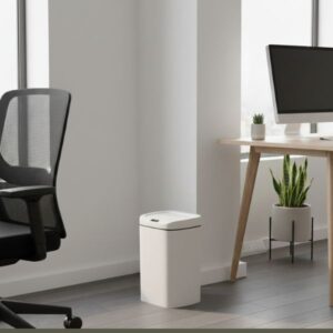 A white smart bin with a dark top is positioned casually in the corner of a modern office. To the right, a light-colored wooden desk holds a computer monitor, a small potted succulent, and a snake plant in a white planter. A black office chair with a mesh back is visible on the left, next to the bin. Large windows are in the background, letting in natural light.