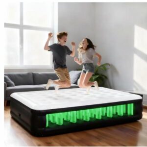 A young boy and girl are shown mid-air, happily jumping on the large inflatable mattress, which has its internal structure colored bright green. The mattress is situated on a wooden floor in a bright room with a grey sofa visible in the background, showcasing the mattress's durability and fun use.