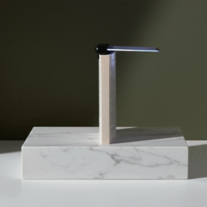A compact, black clip-on LED book light illuminating the pages of a vertically standing paperback book. The book and light are displayed on a white marble rectangular pedestal against a dark green background.