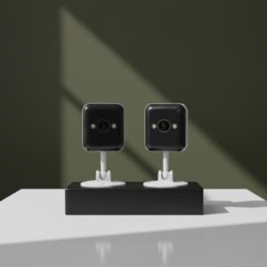 Two white and black security cameras are standing side-by-side on a matte black rectangular block. The background is a dark olive green wall with a prominent soft shadow cast by a window or object.