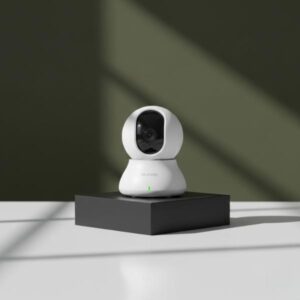 The white dome-shaped camera is standing upright on a matte black rectangular display block, centered on a white surface. The background is a dark olive green wall with a diagonal soft shadow.