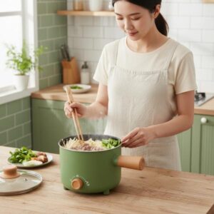 A woman in a white apron stirring food in the green electric cooking pot on a wooden kitchen counter with a light green tiled background.