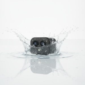 A black mini camera dipping into water, creating a splash and concentric ripples, illustrating its water resistance.