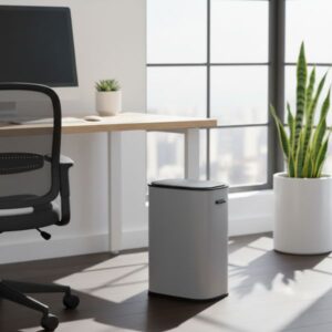 A grey smart bin is placed in a bright, modern office corner. A light wood desk with a computer monitor, a black office chair, and a large potted plant are visible. The background features a large window with a cityscape view. Sunlight streams into the room, creating strong shadows on the floor.