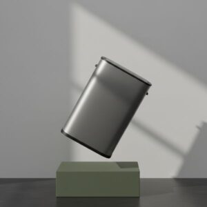 A dark grey smart bin is shown levitating above a matte olive green rectangular block. The bin is tilted at an angle. The background is a light grey wall with strong, sharp shadows cast from a directional light source, emphasizing the product's design.