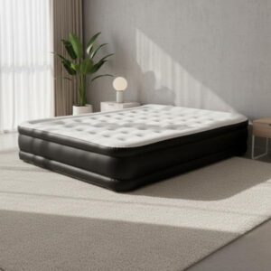 A large, black and white inflatable mattress is placed on a light carpet in a bright, modern room. Sunlight streams in from a nearby window, casting dramatic shadows from the window frame onto the light grey wall. A small table with a lamp is visible next to the mattress.