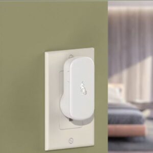 A white doorbell chime (plug-in unit) is inserted into a wall outlet on an olive green wall, with soft lighting suggesting an indoor home setting.