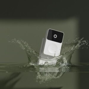 A white smart doorbell unit is shown splashing into water, creating a large crown-shaped splash, demonstrating its water resistance. The lighting is dramatic, casting strong shadows on the water.