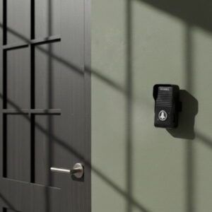 A close-up view of a black doorbell unit installed on an olive green wall next to a modern black door with a silver handle, featuring strong window shadows.