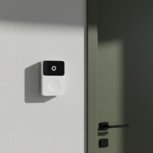 A white rectangular smart doorbell unit is installed on a light grey wall, positioned next to a partially visible dark olive green door with a black handle. Sunlight casts a sharp shadow on the wall