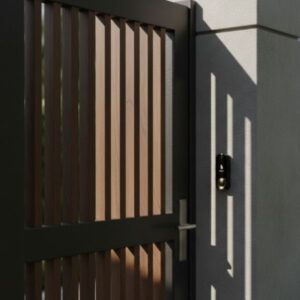 Close-up view of a doorbell unit (appears black/dark) installed next to a modern metal and wood slat main gate, with strong shadows cast by the gate structure.
