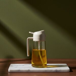 A clear glass oil bottle featuring a light-colored (white/cream) automatic gravity pouring spout, resting on a white marble display block.