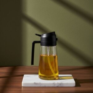 A clear glass oil bottle equipped with a dark (black) automatic gravity pouring spout and handle, presented on a white marble slab.