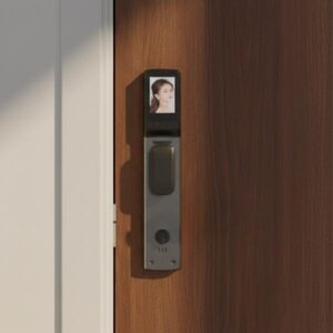 : A vertical, dark grey smart door lock is mounted on a light brown wooden door. Sunlight shines from the side, illuminating the lock and casting a shadow on the door. The screen displays a person's face.