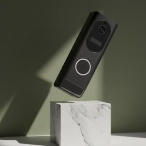 A black rectangular doorbell unit is shown levitating above a white marble block with green veins. The doorbell is tilted, and the background is an olive green wall with a prominent shadow.