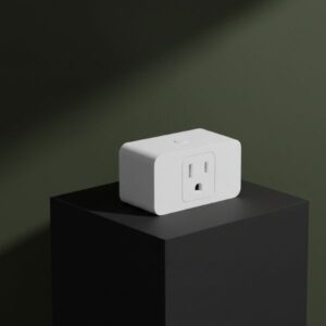 A white rectangular smart plug, featuring a two-pronged outlet and a small button on top, rests aesthetically on a matte black block. The background is a dark olive green with soft, natural shadows.