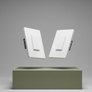 Two white smart light switches are levitating in mid-air, angled away from each other, above a matte olive green block. The background is a soft gray.